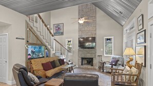 A spacious living room with a stone fireplace, comfortable seating, a television, and a ceiling fan.