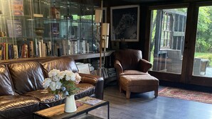 A modern living room with a leather sofa, a coffee table with a vase of flowers, a bookshelf, and a large window with a view of greenery. - Cozy designer Cottage on private historic estate-near NYC-Oasis in Metropolis (West Caldwell)