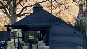 A small, dark-colored cabin with a sloped roof, a porch with wicker furniture, and a stone fireplace. - Cozy designer Cottage on private historic estate-near NYC-Oasis in Metropolis (West Caldwell)