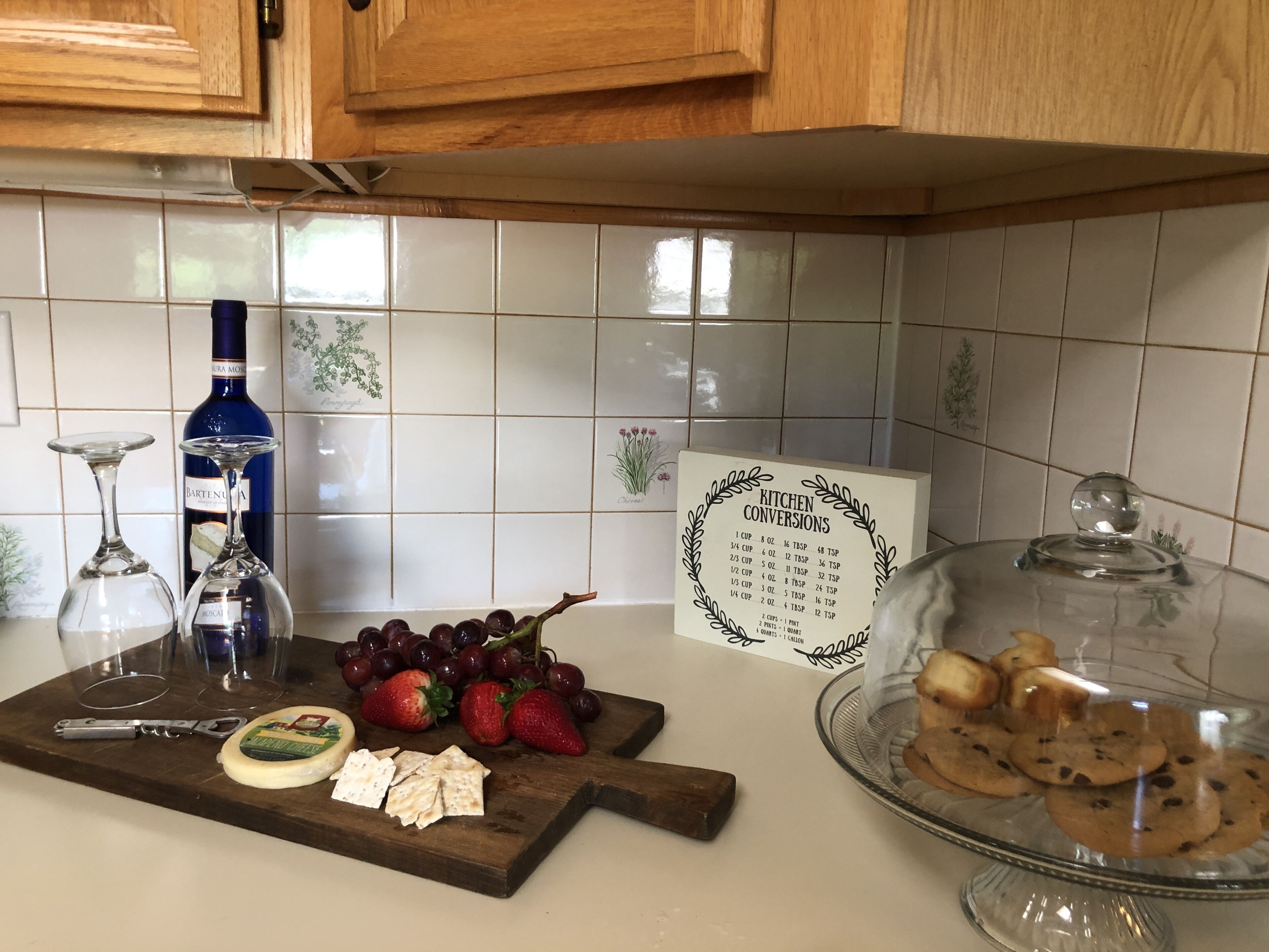 A kitchen counter with a wine bottle, glasses, a fruit platter, a cheese board, and a cookie display.