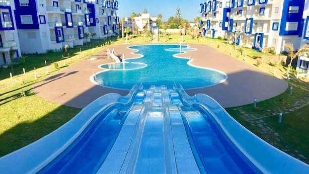 A water slide leading to a pool surrounded by modern buildings with balconies. - Great Pool View Apartment in Kariat Cabo 4548 (Cabo Negro)