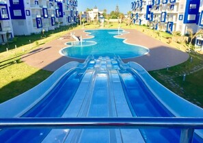 A water slide leading to a pool surrounded by modern buildings with balconies. - Great Pool View Apartment in Kariat Cabo 4548 (Cabo Negro)