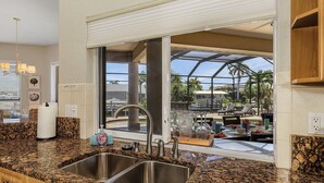 Une cuisine dotée de plans de travail en granit, d’éviers en acier inoxydable et offrant une vue sur un patio extérieur aménagé avec des sièges et une structure en verre.