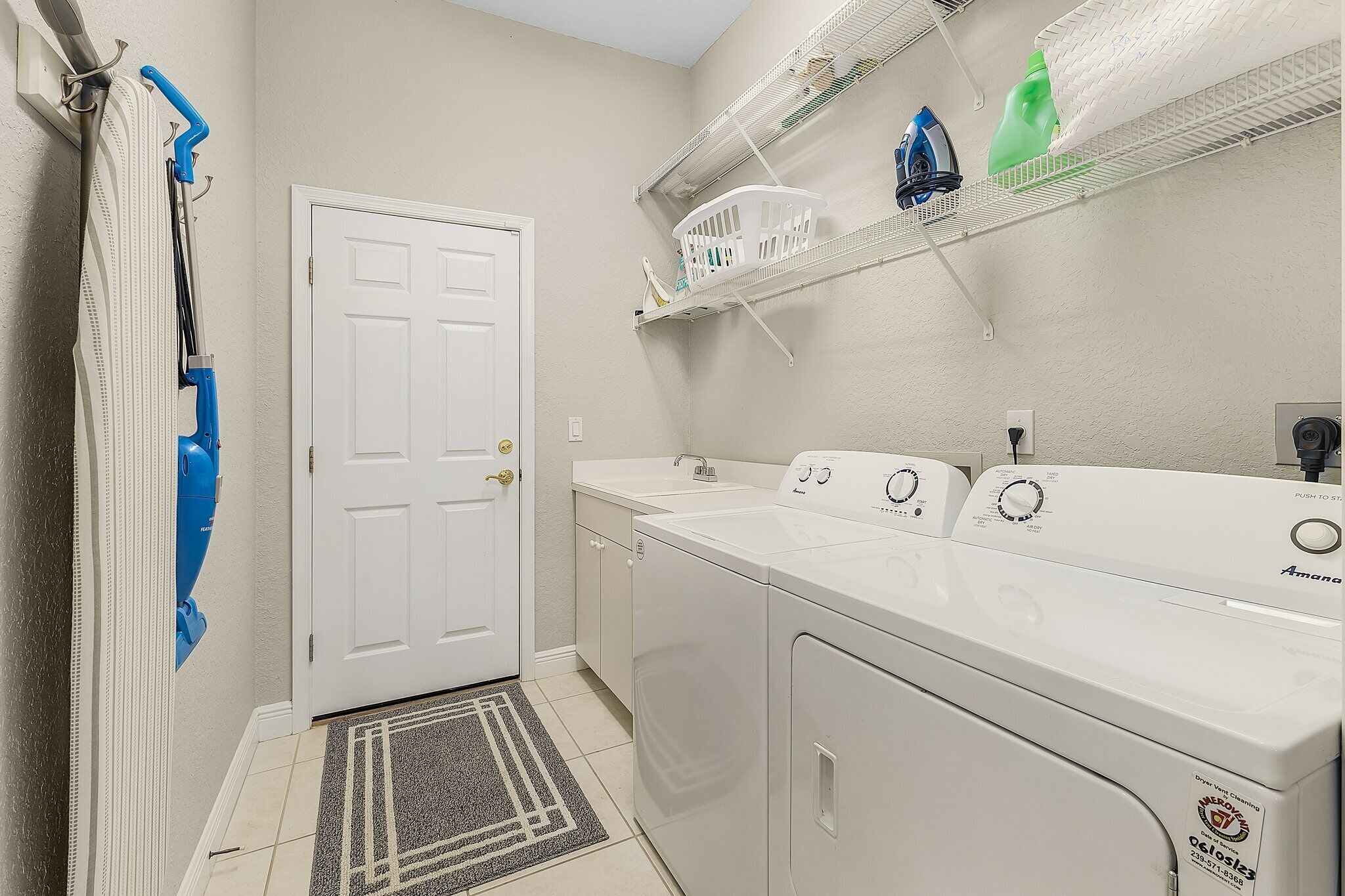A laundry room with a front-loading washer and dryer, a white door, a small rug, and shelves above holding laundry supplies and a green bottle.
