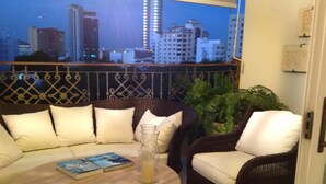 A balcony with wicker furniture, a round table with a book and snacks, and a city view at dusk.