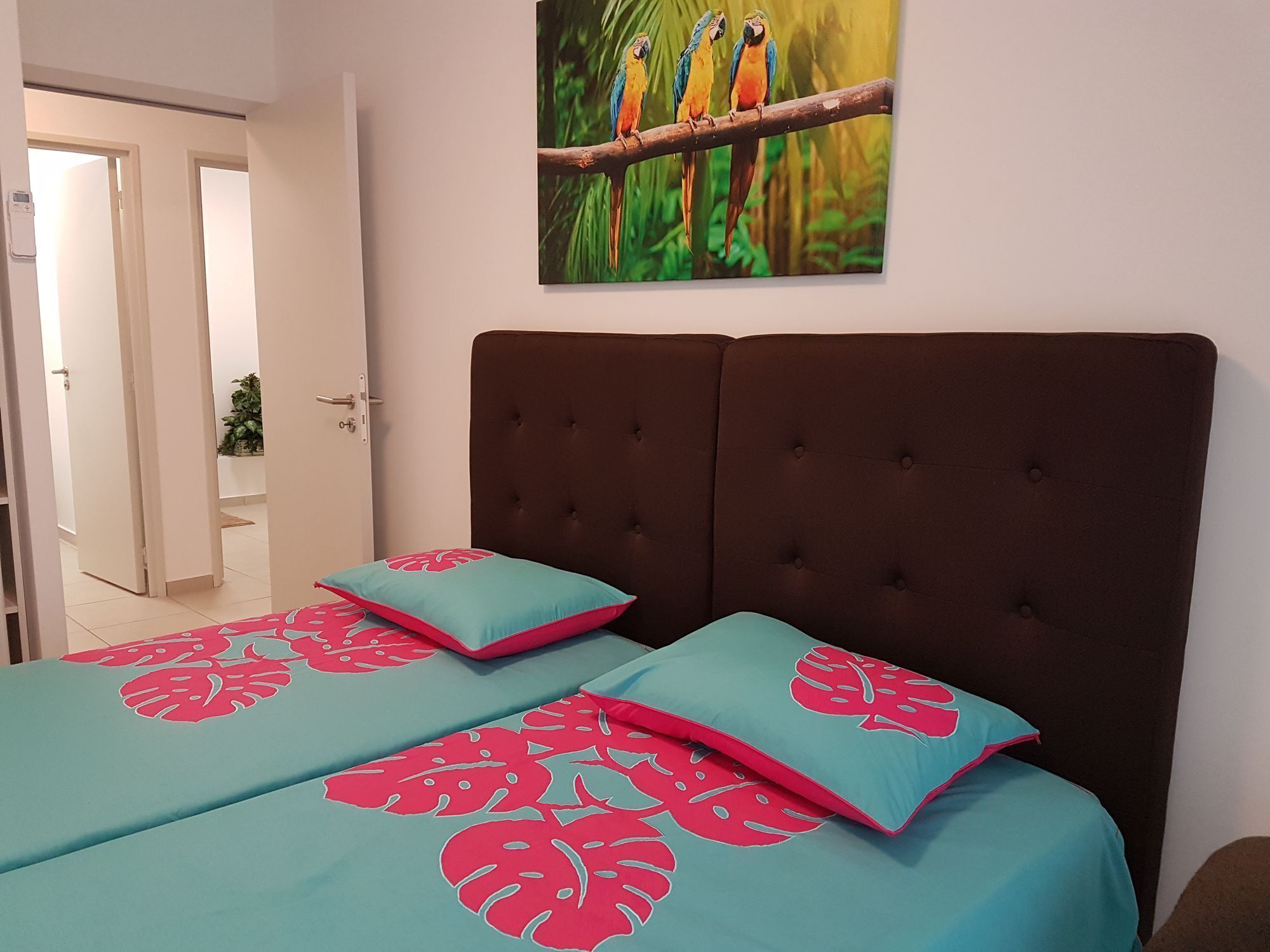 A bedroom with a large bed, two pillows with a tropical leaf pattern, a dark headboard, and a painting of colorful birds on the wall.