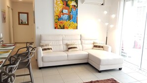 Un salon avec un canapé d’angle blanc, une table à manger avec des chaises, un tableau au-dessus du canapé et une grande fenêtre avec des stores.