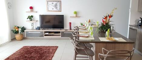 A modern living room with a flat-screen TV, a dining area with a glass table, and a kitchen area with cabinets.