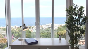 A desk with a lamp and a book, a potted plant, and a view of the sea and buildings through large windows.