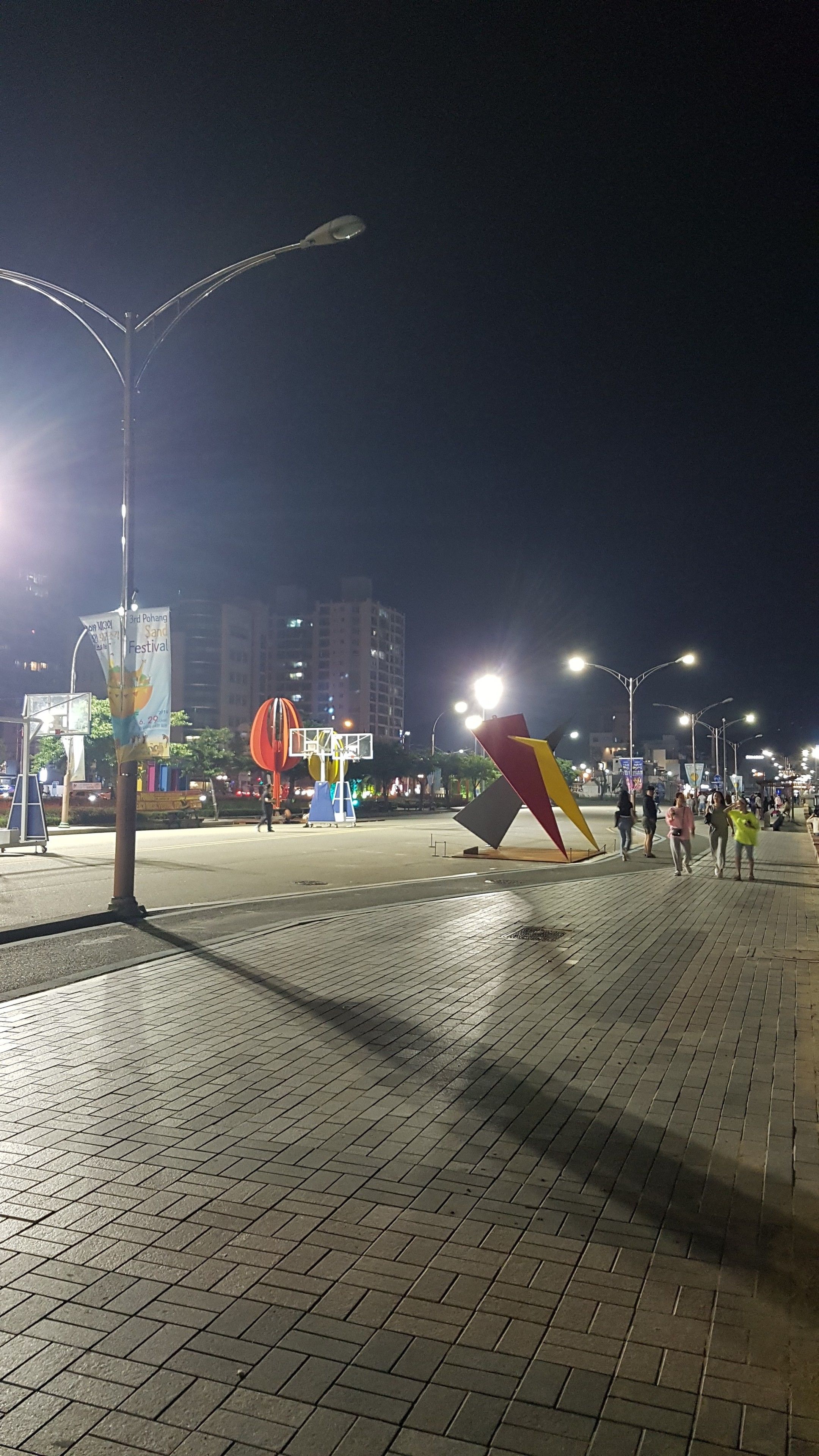 A nighttime urban street scene with a paved walkway, street lamps, and a large colorful sculpture.