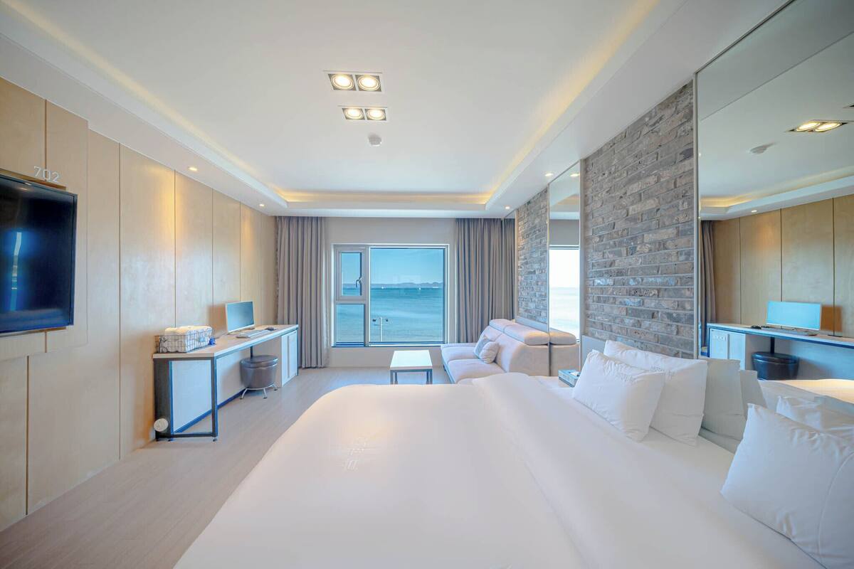 A modern hotel room with a large bed, a desk, a flat-screen TV, and a view of the sea.