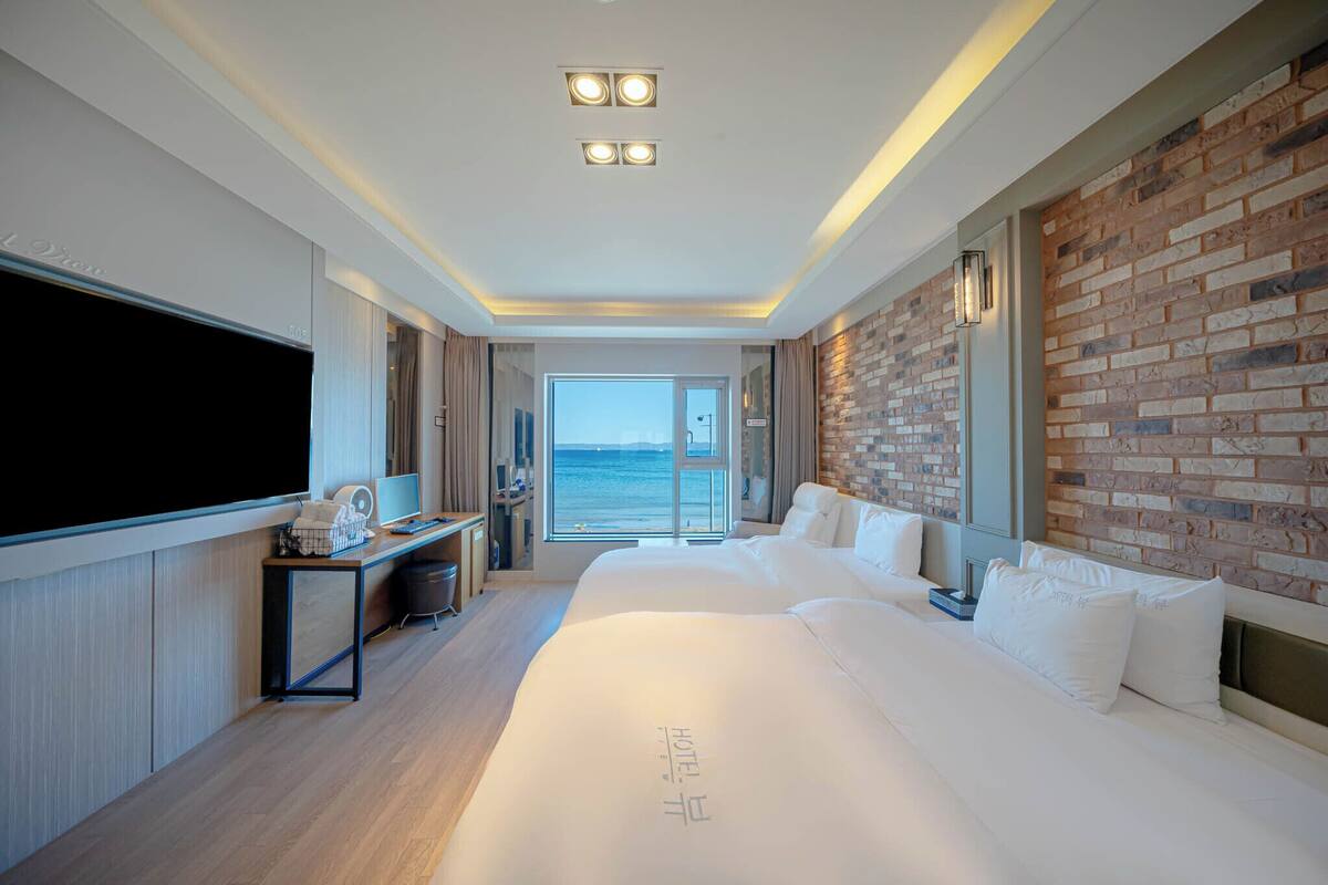 A modern hotel room with a large bed, a flat-screen TV, a desk with a computer, and a brick accent wall.