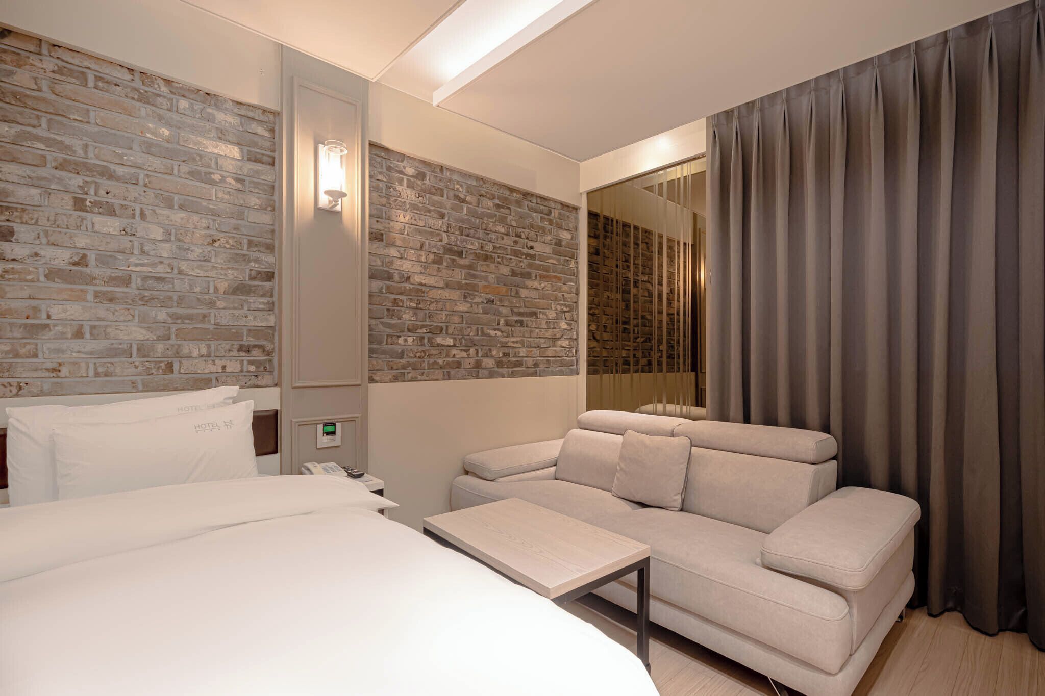 A hotel room with a brick wall, a sofa, a bed, a small table, and a curtain.