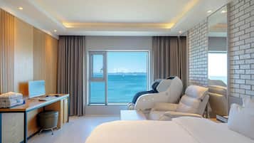 A modern hotel room with a large bed, a desk with a computer, a massage chair, and a view of the sea.
