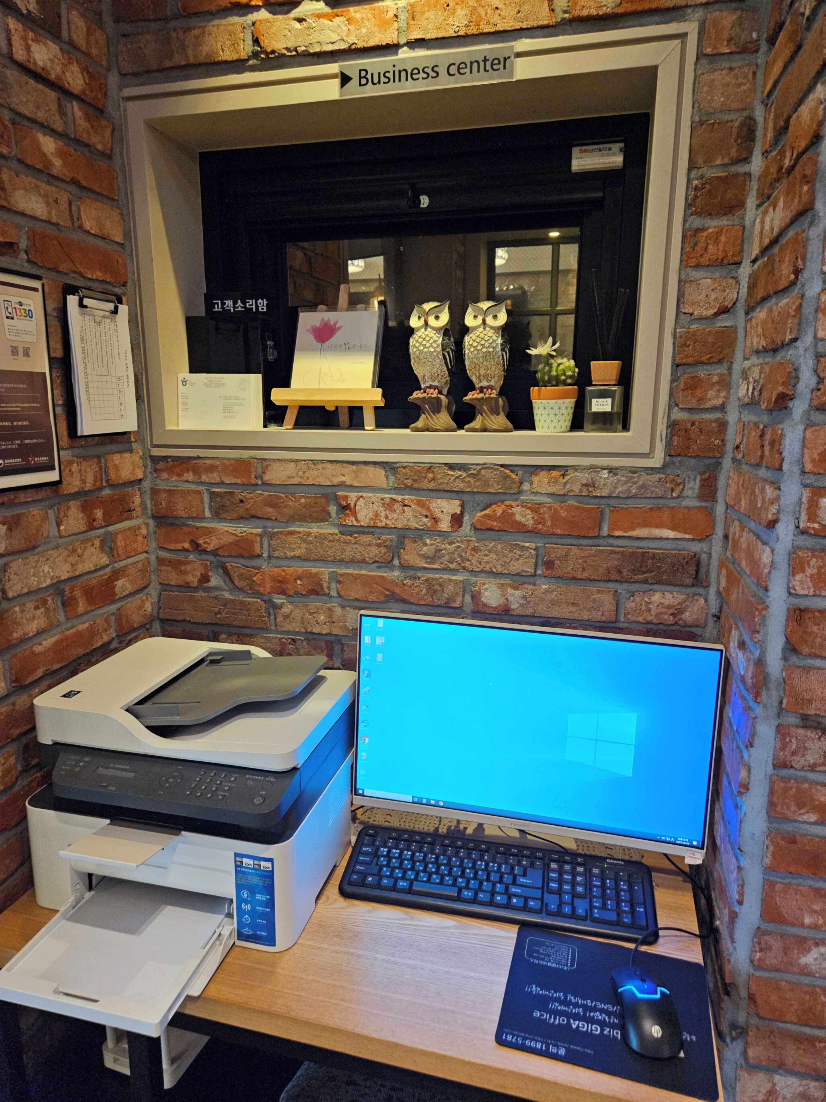 A workspace with a computer, printer, and keyboard, a window with a view of a business center, and a brick wall.