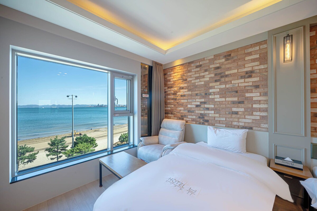 A hotel room with a large window offering a view of the beach and sea, a bed with a 'HOTEL' sign, a brick accent wall, and a white chair.