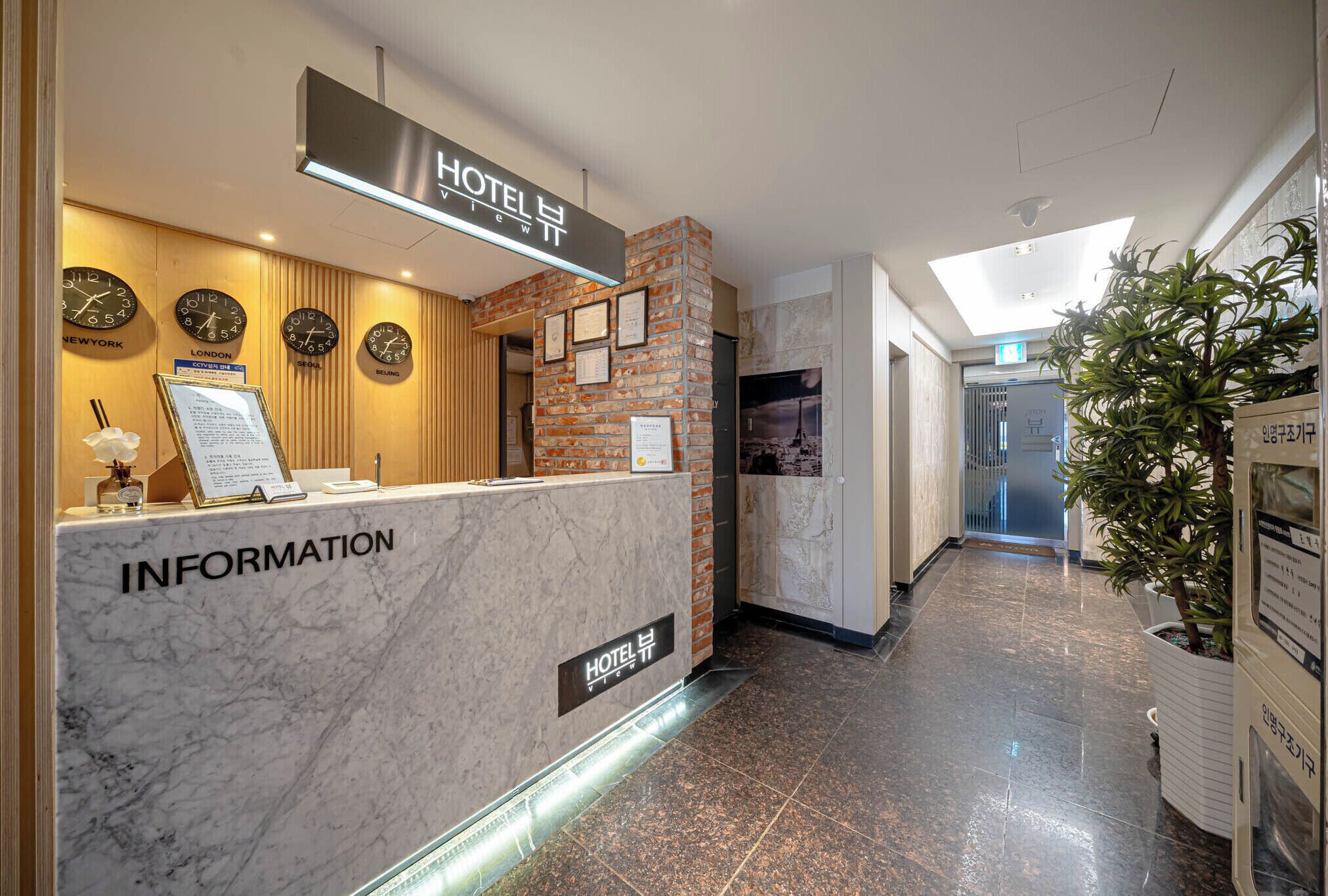 A hotel reception area with a marble counter labeled "INFORMATION," clocks displaying different cities, and a brick accent wall.