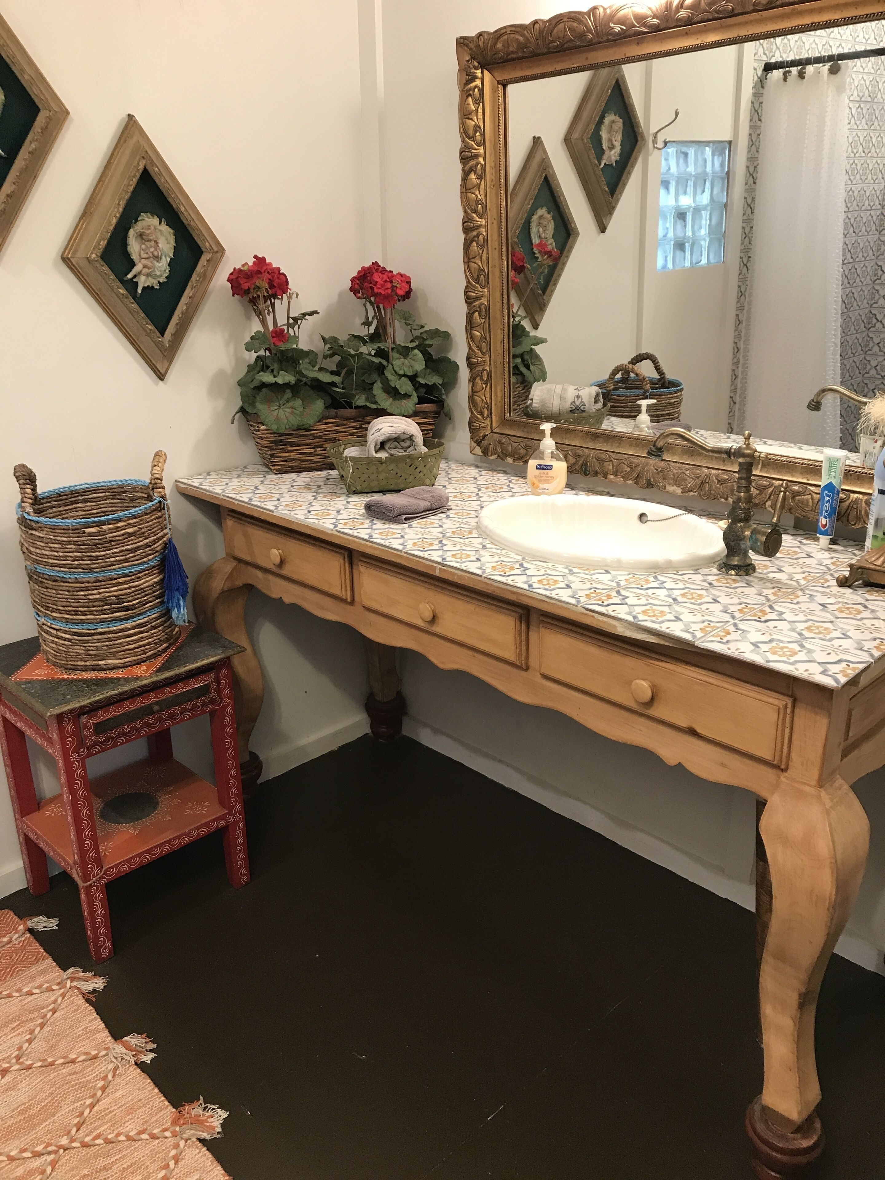 A bathroom with a large mirror, a white sink, and a wooden vanity. There are decorative baskets and a small stool.
