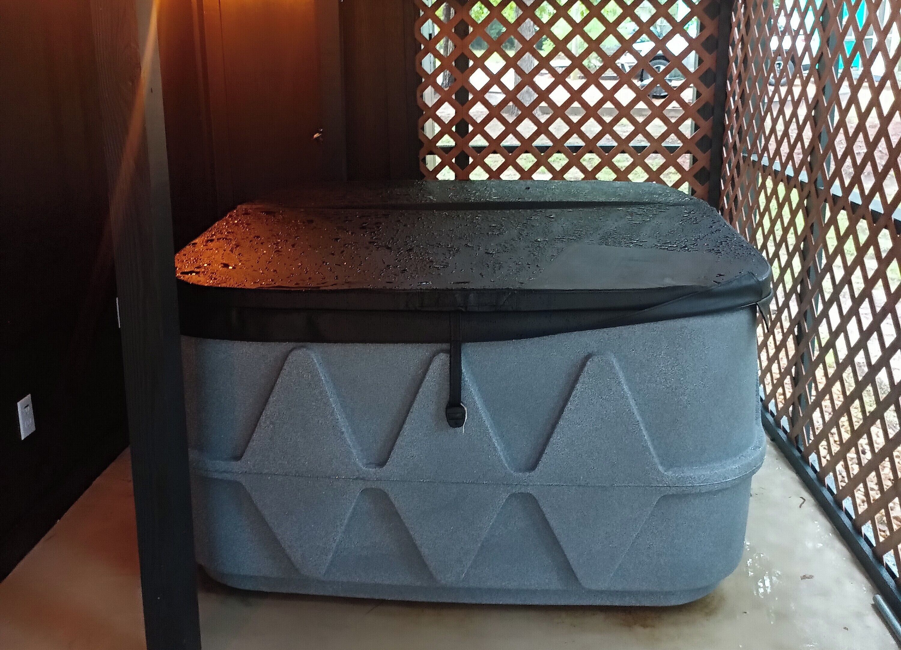 A large, grey, rectangular storage bin with a textured surface and a black top, placed outdoors near a lattice fence.