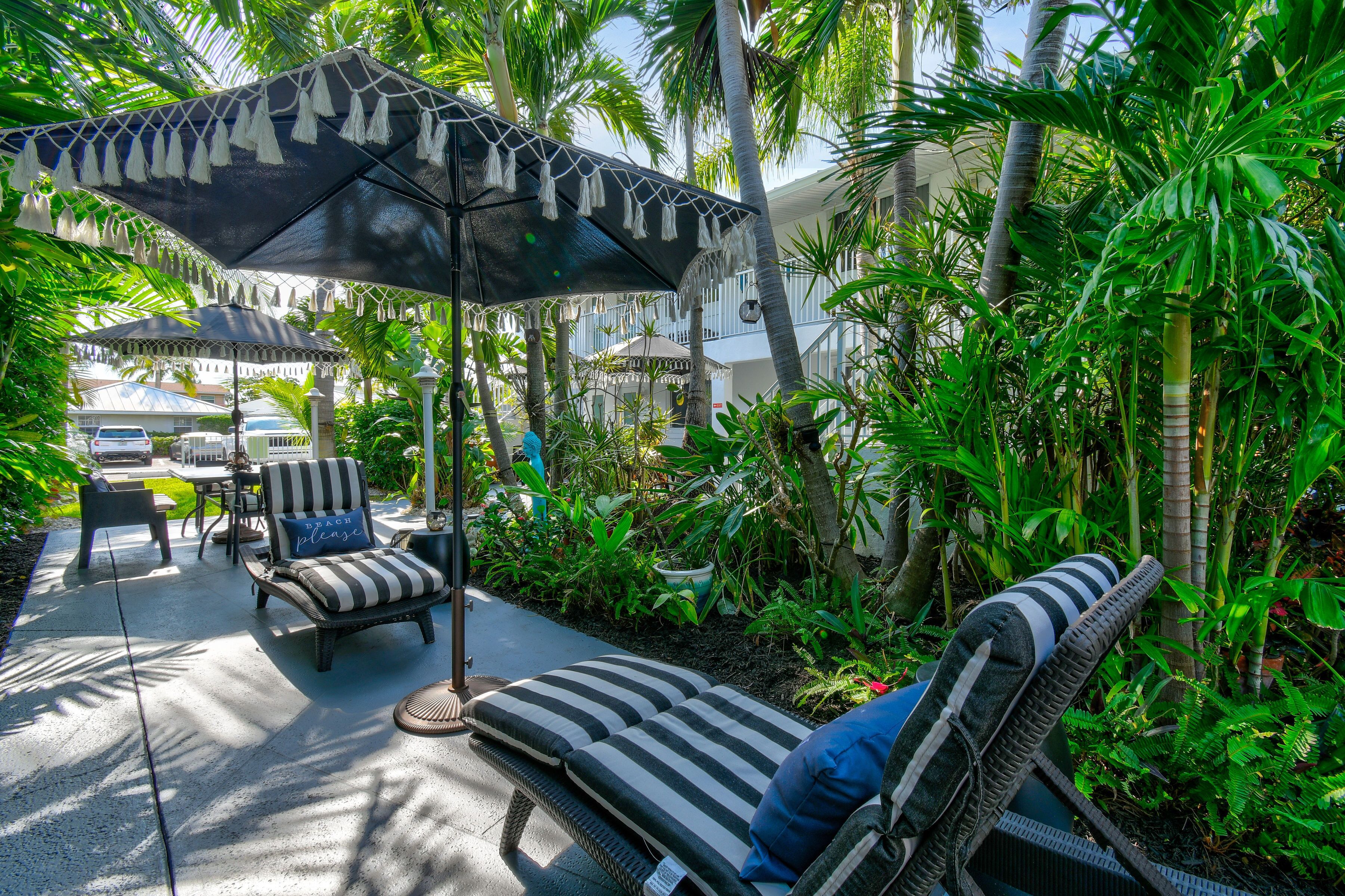 Un espace extérieur aménagé dans un style tropical, avec des fauteuils de jardin à rayures, un parasol et une végétation luxuriante.