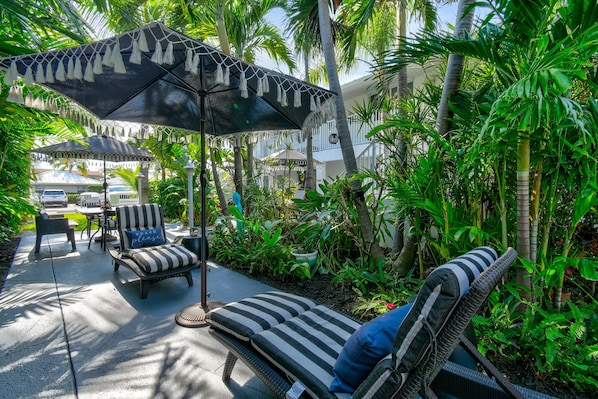 A tropical outdoor seating area with striped lounge chairs, a covered umbrella, and lush greenery. - one bedroom furnished apartment by beach (West Palm Beach)
