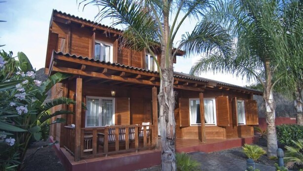 A wooden two-story house with a balcony, surrounded by tropical plants and palm trees.
