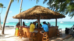 Eine Holzterrasse mit einem Reetdach, umgeben von Palmen, mit Blick auf einen Strand mit klarem türkisblauem Wasser und Booten in der Ferne.