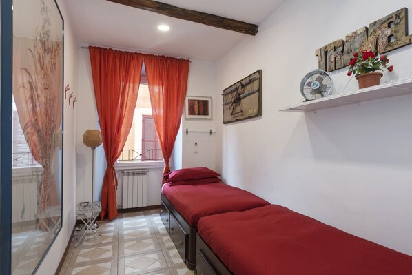 A small room with a single bed, a window with orange curtains, a wall-mounted fan, and a shelf with a potted plant.