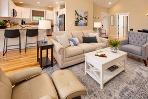 A modern living room with a beige sofa, a white coffee table, and a grey armchair. There is a kitchen in the background with white cabinets and a refrigerator. - Family-Run Cottage with Fireplace, 2 Miles to Asheville (Asheville)