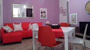 A living room with a red sofa, white table, and red chairs. There is a mirror on the wall and a clock. - CASE COLOMBO AND MATTARELLA central apartment with wi-fi and free parking space (San Vito Lo Capo)