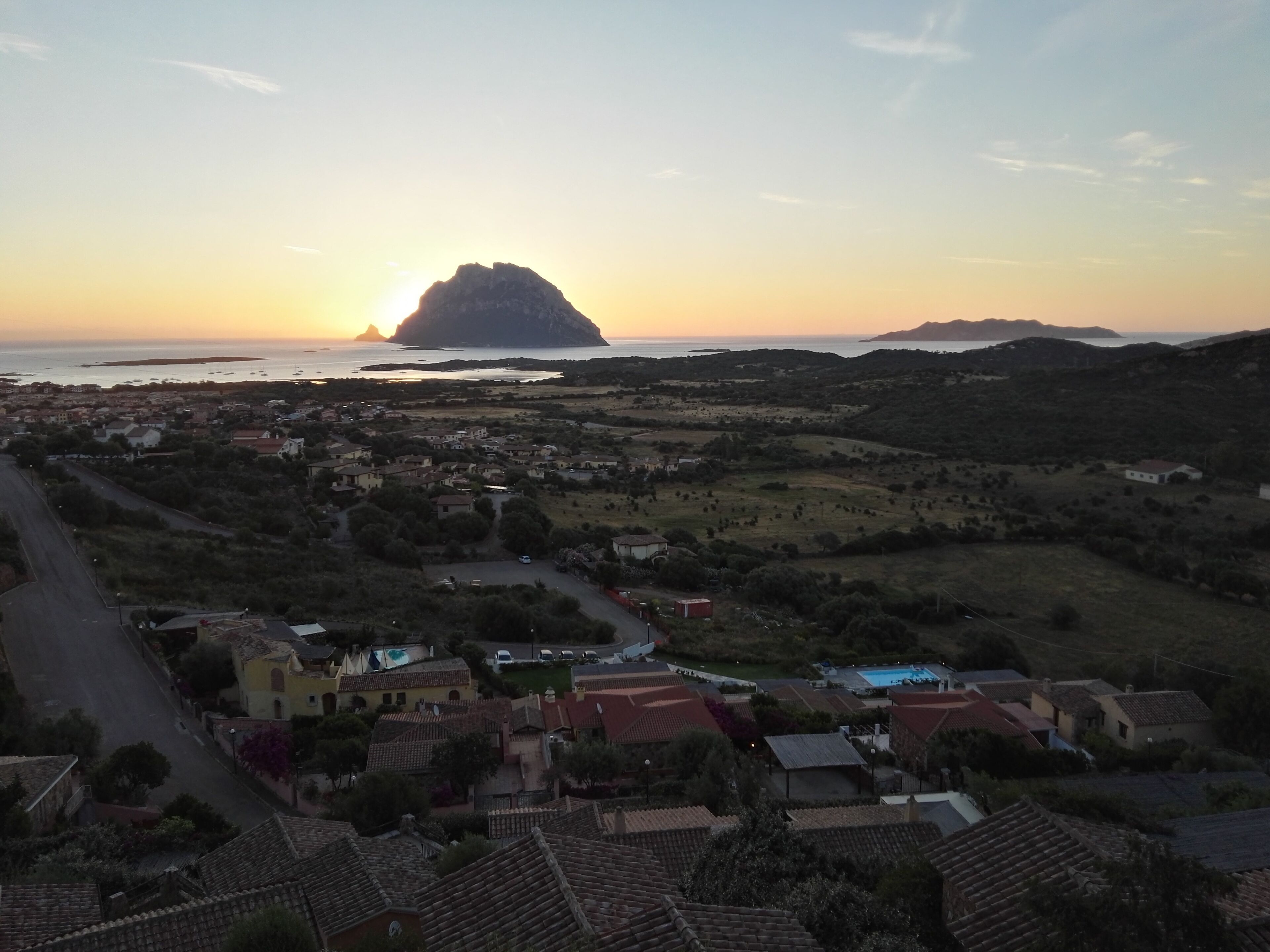 A coastal town with terracotta-roofed buildings, a central street, and a view of the sea and a prominent hill in the distance.