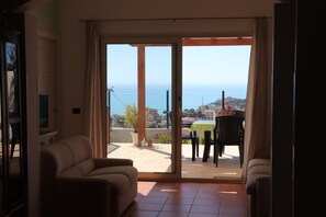 Een woonkamer met een schuifdeur die leidt naar een balkon met een tafel en stoelen, met uitzicht op een schilderachtig zeegezicht en gebouwen.