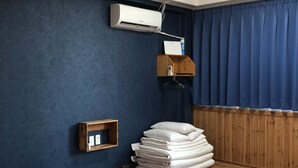 Un dormitorio con suelo de madera, una cama con colcha blanca, una pila de toallas dobladas, un equipo de aire acondicionado y un estante de madera con un objeto pequeño.