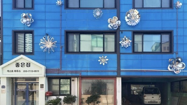 Un edificio azul con la palabra "Good" en la fachada, flores de metal decorativas, entrada de una casa de huéspedes y un coche aparcado en el garaje.