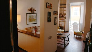 Een woonkamer met een bank, een tafel en een boekenkast. Een deur leidt naar een andere kamer.