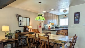 A dining area with a wooden table and chairs, a hanging green light fixture, and a kitchen in the background.