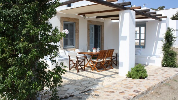 A covered outdoor seating area with a table and chairs, a stone floor, and a white building with blue shutters. - Chora villa 1 with unique sea view (Hora)