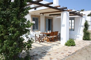 A covered outdoor seating area with a table and chairs, a stone floor, and a white building with blue shutters. - Chora villa 1 with unique sea view (Hora)