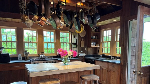 A kitchen with wooden cabinets, a large island with stools, hanging pots, and a view of greenery through the windows. - Bright and cozy menemsha cottage (Chilmark)