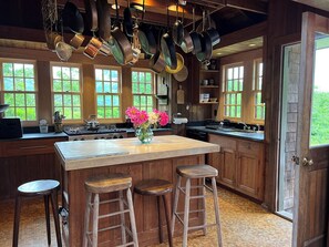 A kitchen with wooden cabinets, a large island with stools, hanging pots, and a view of greenery through the windows. - Bright and cozy menemsha cottage (Chilmark)