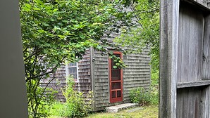 A wooden deck leading to a red door, surrounded by a grassy area and a stone pathway. - Bright and cozy menemsha cottage (Chilmark)