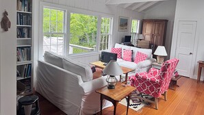 A cozy living room with a white sofa, a red patterned armchair, and a wooden coffee table. There is a bookshelf filled with books and a lamp on the table. - Bright and cozy menemsha cottage (Chilmark)