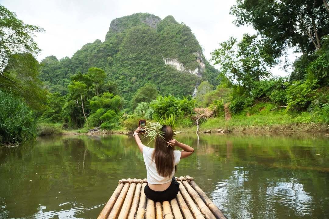 A person sitting on a bamboo raft in a river surrounded by lush greenery and a mountain in the background.