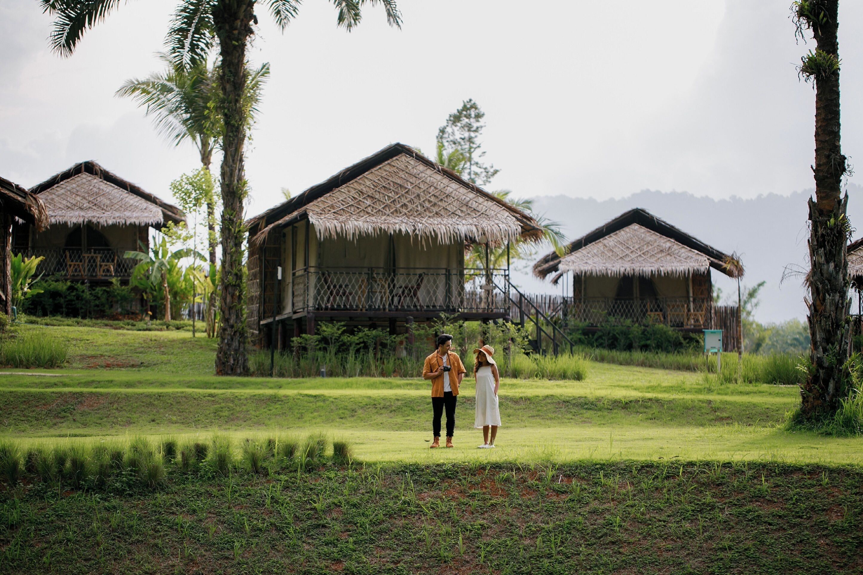 A couple walking on a grassy area with thatched-roof bungalows in the background.