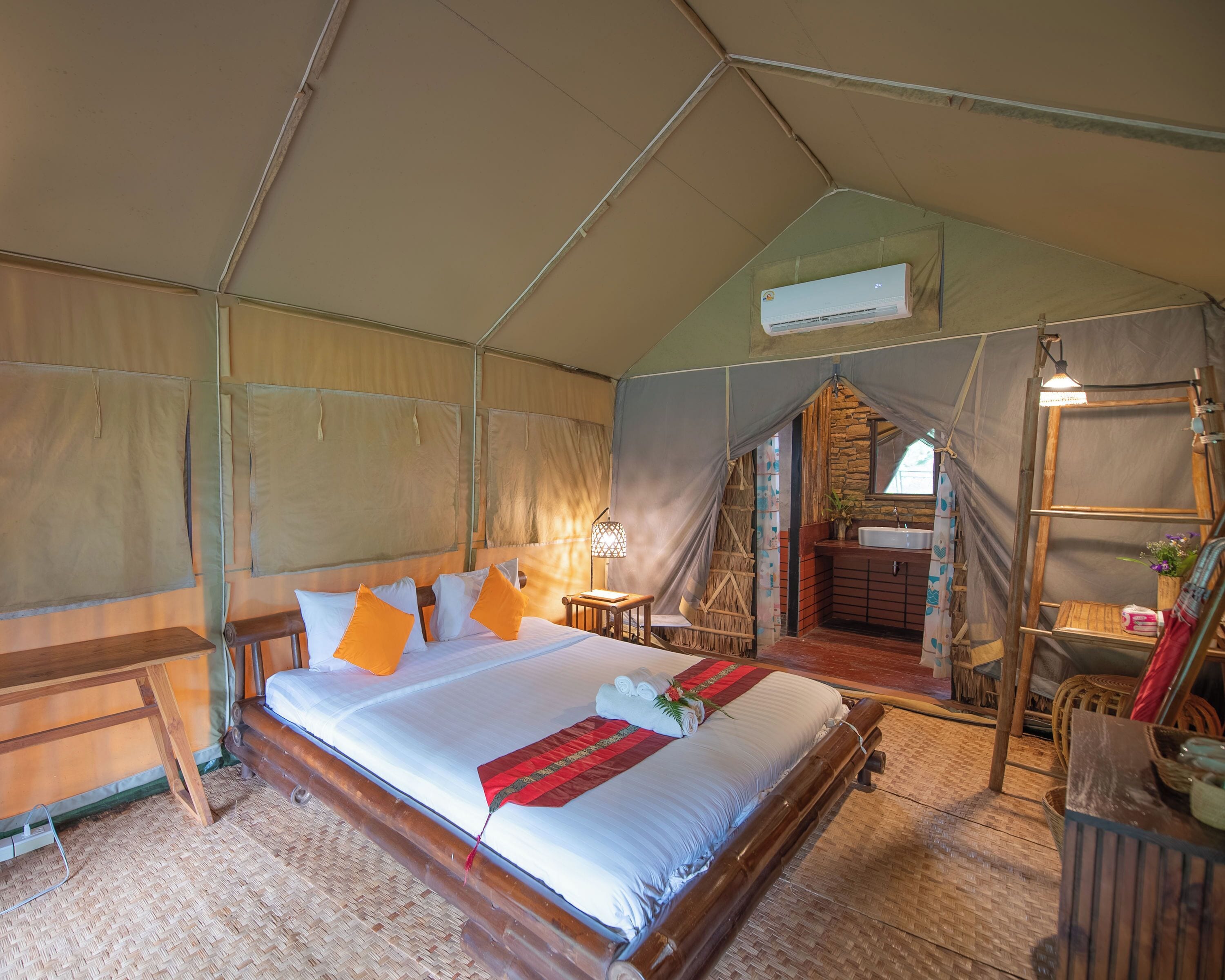 Habitación tipo alojamiento en una tienda de campaña, con una cama, una mesita, una lámpara, una escalera de madera y un baño visible a través de una puerta.