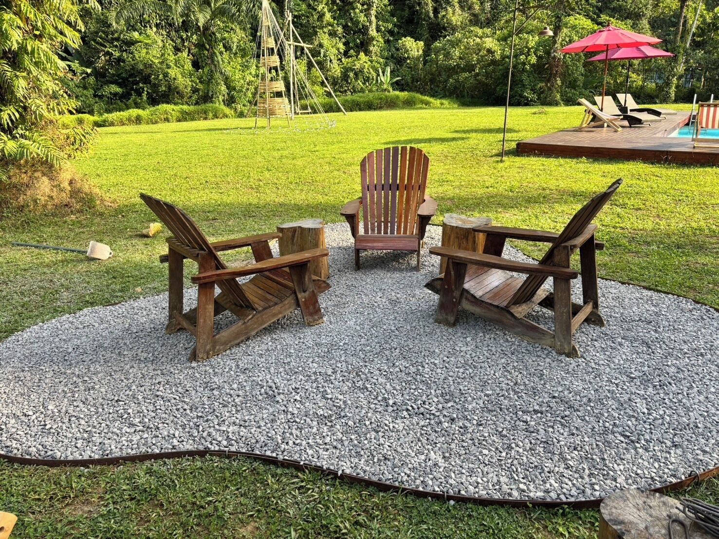 A circular seating area with Adirondack chairs and a wooden table, surrounded by a gravel border, in a lush garden setting.
