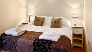 A neatly made bed with two pillows, a bedside table with a lamp, and a small drawer unit.