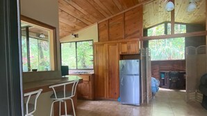A wooden interior with a kitchen area, bar stools, a refrigerator, and a window with a view of greenery.