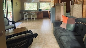 A spacious living area with a wooden ceiling, a sofa, a dining table, and a kitchen area with a view of greenery outside.