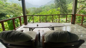 A covered outdoor seating area with two cushioned chairs and a table, overlooking a lush, mountainous landscape.